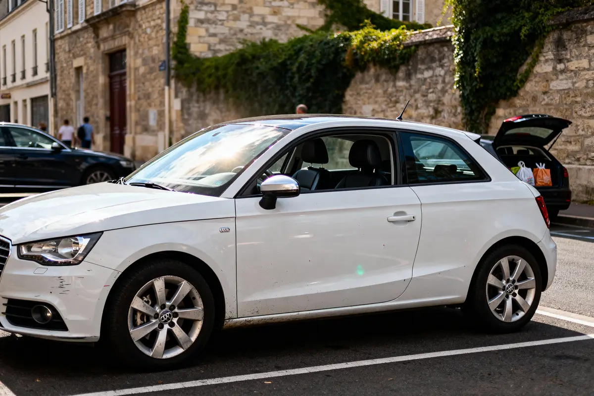Voiture blanche garée en ville, bâtiment en pierre et hayon ouvert en arrière-plan.