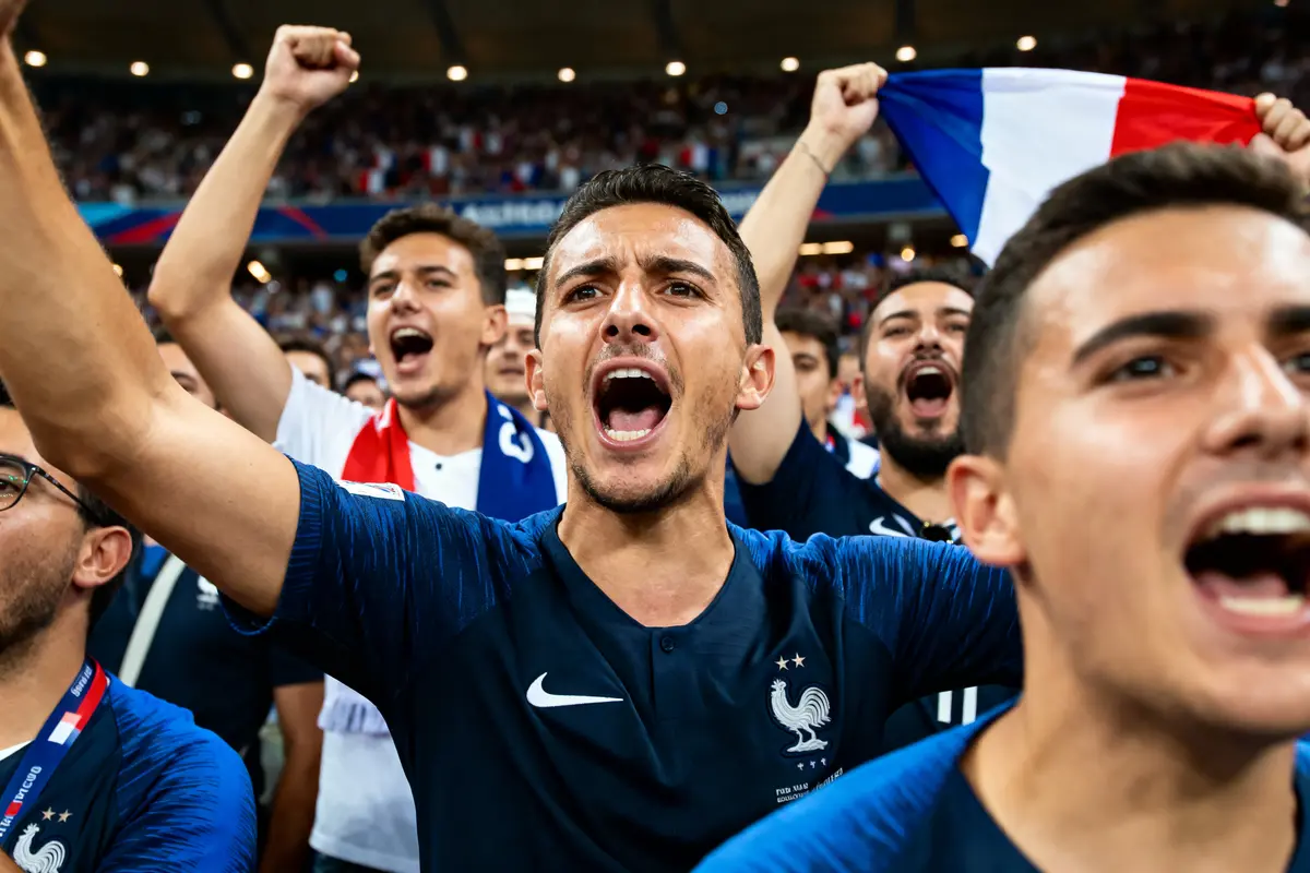 Supporters français enthousiastes, criant avec drapeau tricolore, lors d'un match de football, arborant maillots bleu, blanc, rouge.