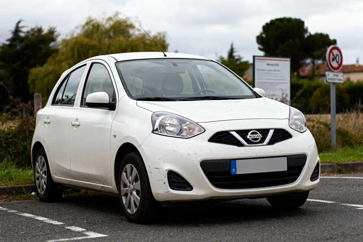 Voiture blanche Nissan garée sur un parking, avec arbres et panneaux en arrière-plan, ciel nuageux.