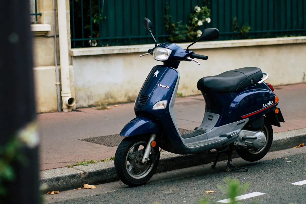 Scooter bleu Peugeot Ludix garé sur le trottoir, à côté d'une clôture verte et d'un mur beige.