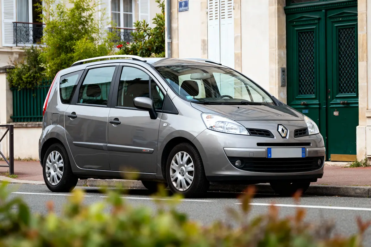 Voiture Renault grise garée devant un bâtiment résidentiel avec fenêtres encadrées de volets blancs.