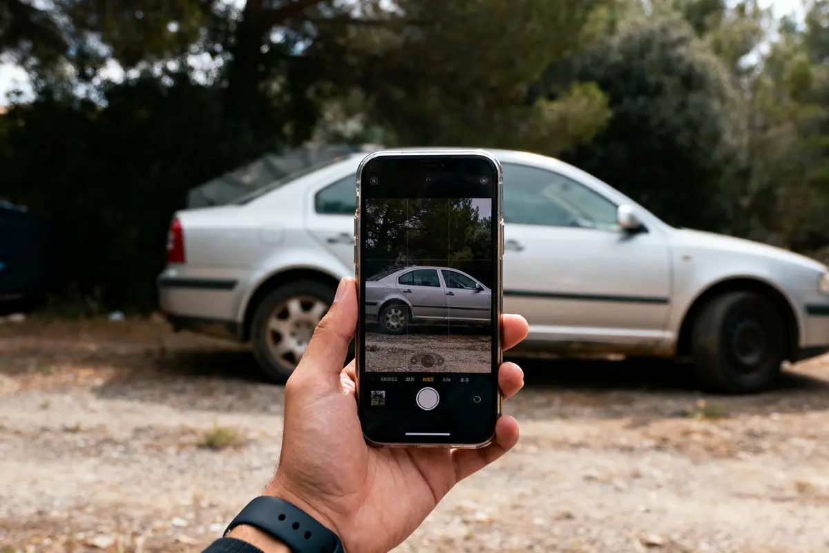 Main tenant un smartphone photographiant une voiture blanche garée devant des arbres dans un parking en plein air.