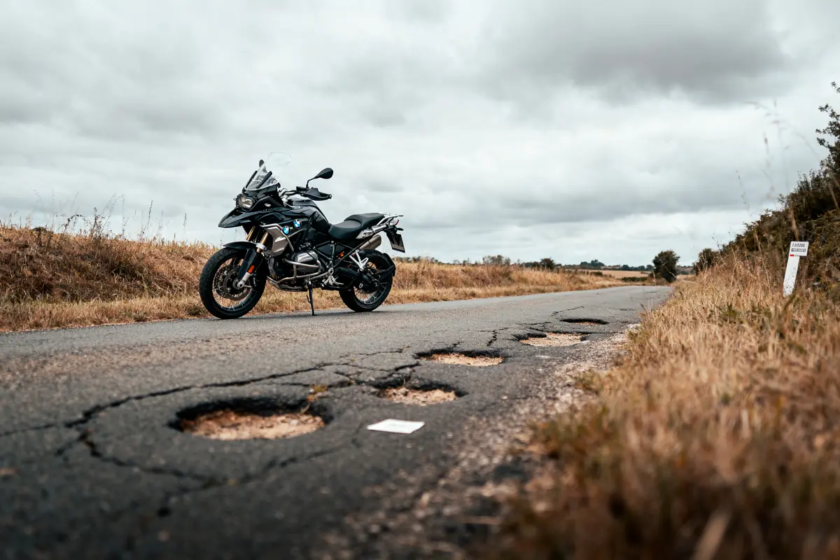 Moto noire garée sur route abîmée par nids-de-poule, ciel nuageux, paysage rural environnant.