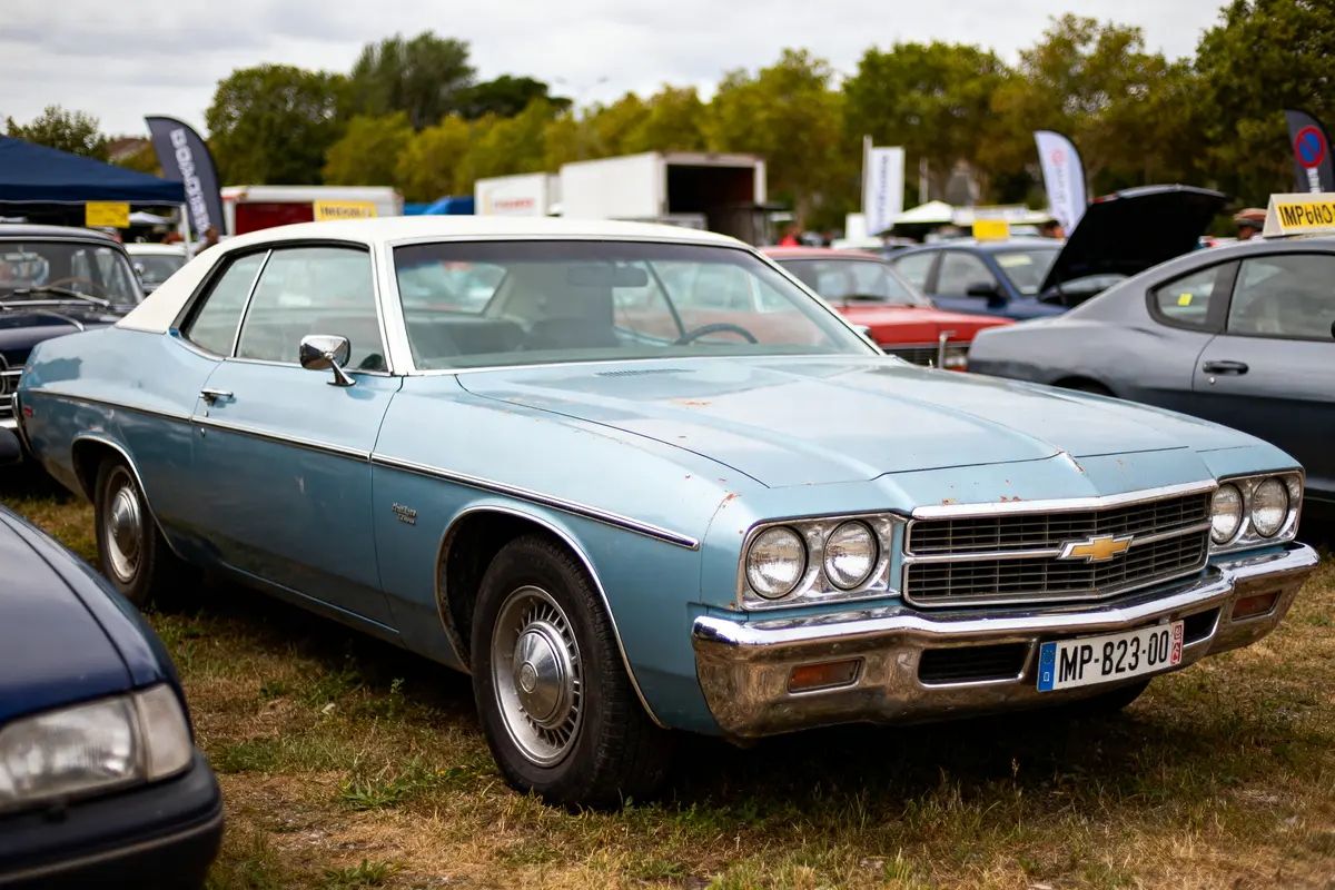 Voiture classique bleue Chevrolet stationnée dans un parc automobile, entourée d'autres véhicules, journée nuageuse.