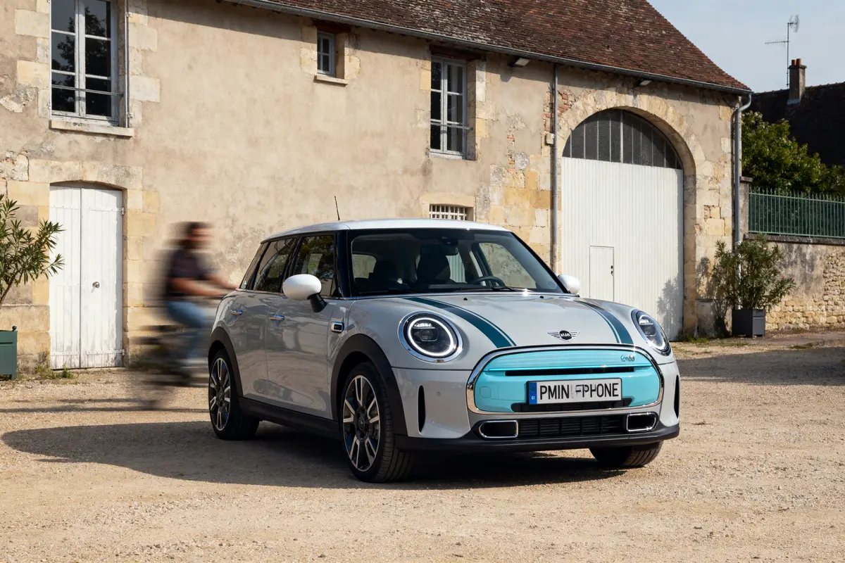 Voiture électrique Mini stationnée devant bâtiment en pierre, cycliste flou passant à proximité.