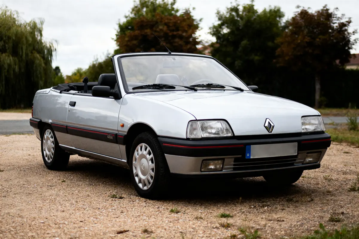 Voiture décapotable grise Renault, stationnée à l'extérieur, entourée d'arbres et de végétation.
