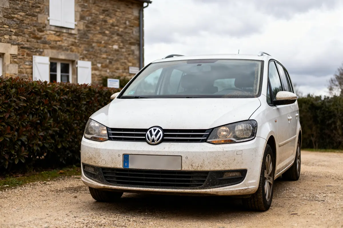 Voiture Volkswagen blanche sale stationnée devant maison en pierres, ciel nuageux en arrière-plan.