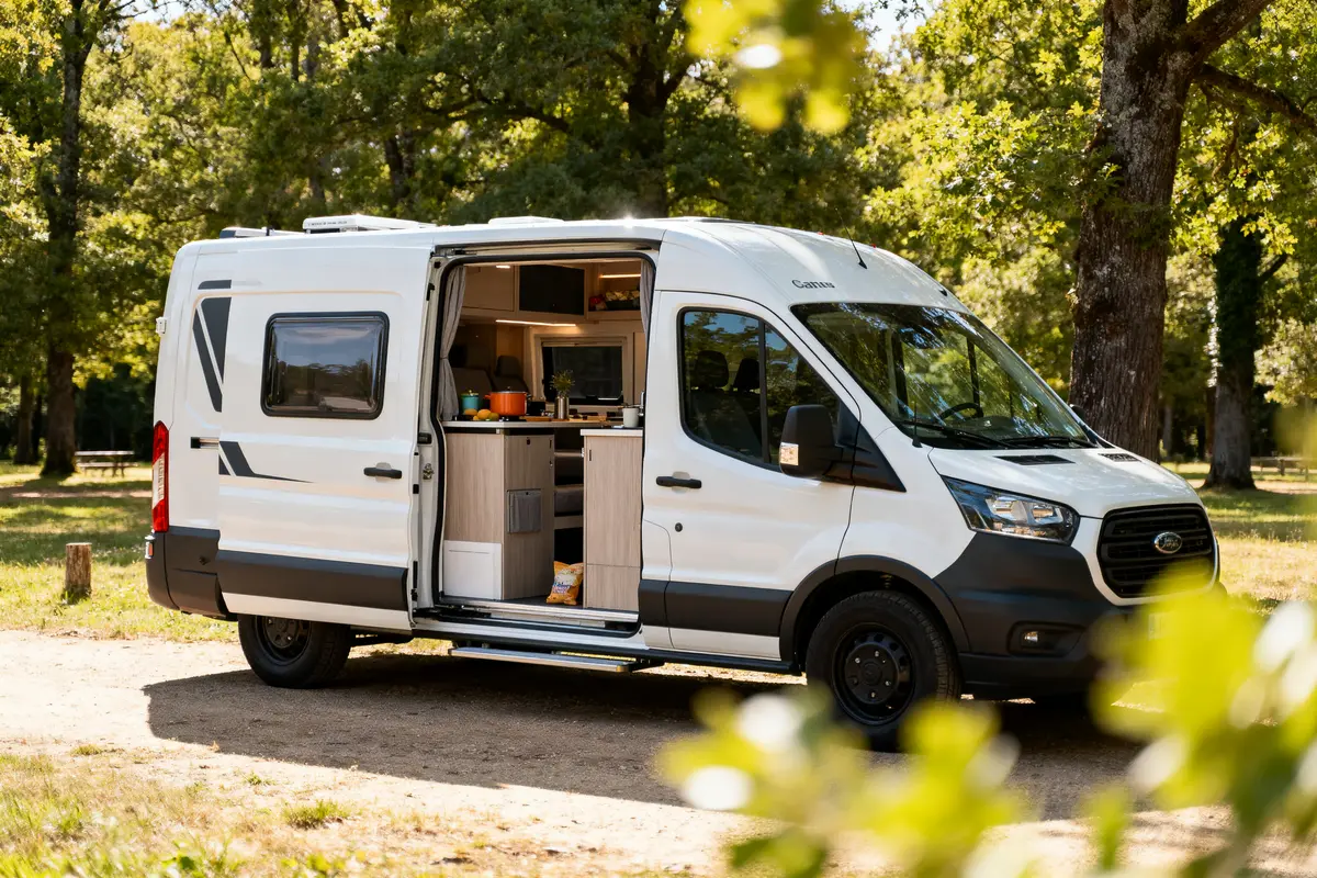 Fourgon aménagé blanc avec porte ouverte, cuisine intérieure visible, stationné dans un cadre forestier verdoyant.