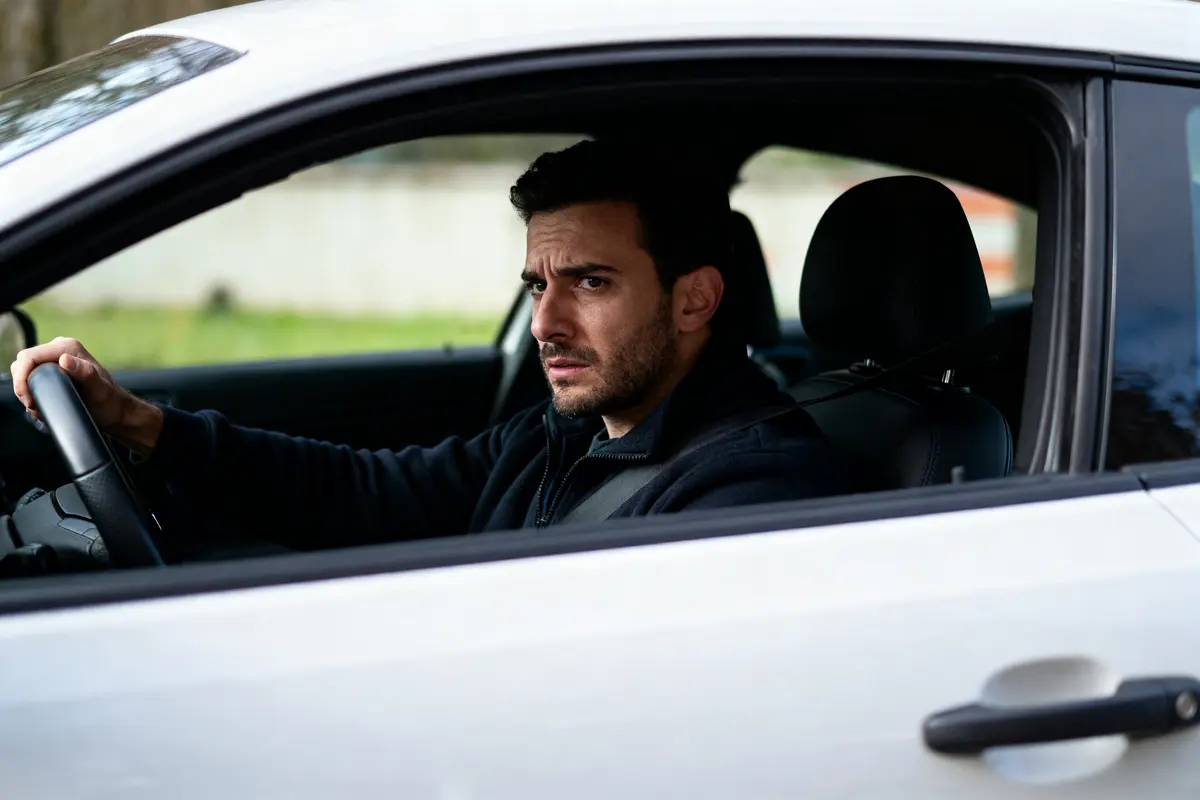 Homme à l'air préoccupé conduisant une voiture blanche, regardant par la fenêtre.