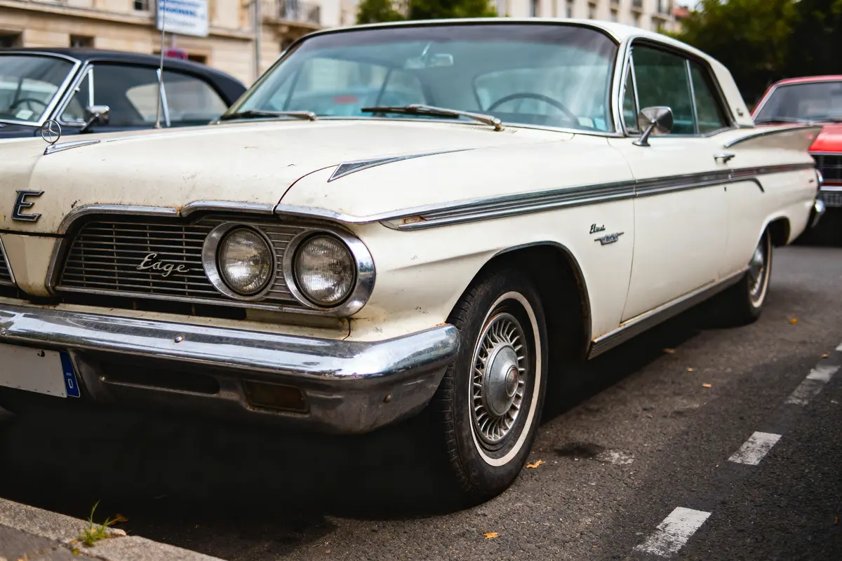 Voiture classique blanche ancienne garée dans la rue, avant avec phares et calandre visibles.