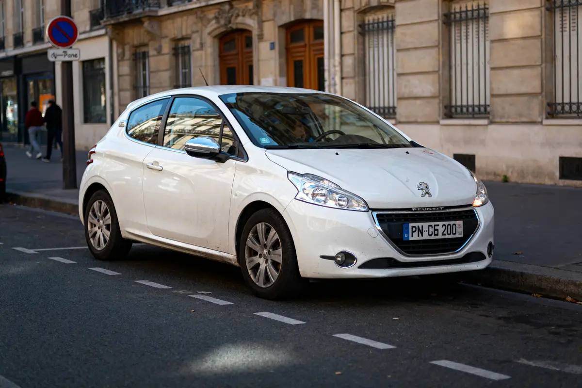 Voiture blanche Peugeot garée en ville, rue bordée d'immeubles historiques et piétons. Plaque PN-CG 200.