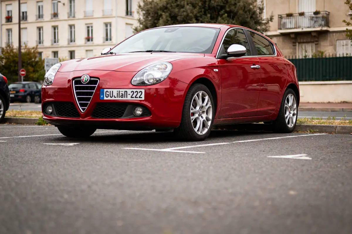 Voiture rouge Alfa Romeo Giulietta stationnée dans un parking urbain, bâtiments en arrière-plan.