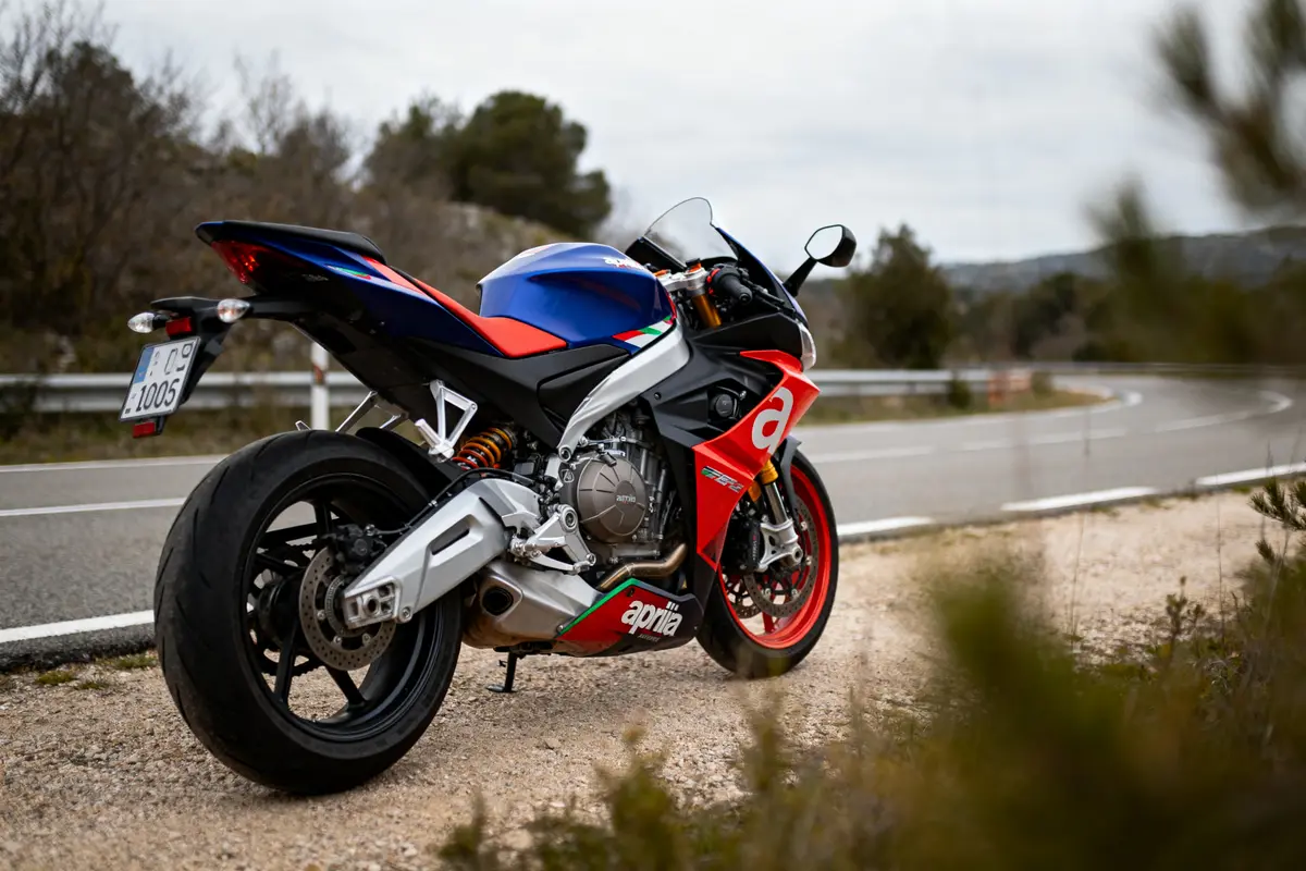 Moto sportive Aprilia colorée garée sur route de campagne sinueuse avec horizon boisé.
