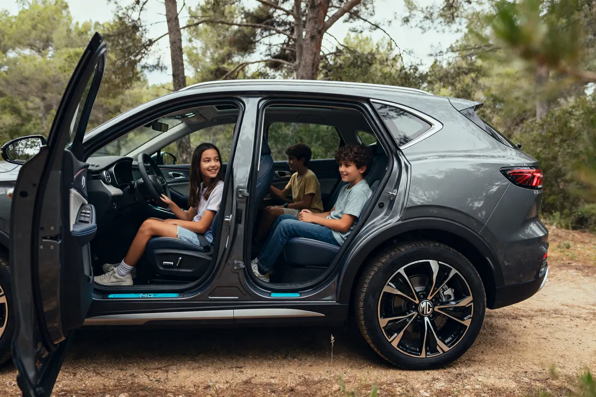 Trois enfants souriants assis dans une voiture grise avec portes ouvertes, entourés d'arbres.