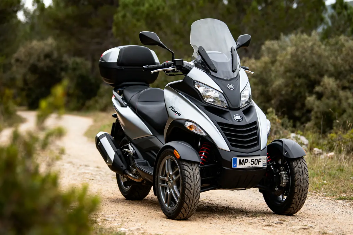 Scooter à trois roues sur chemin forestier, couleur grise et noire, top-case arrière.
