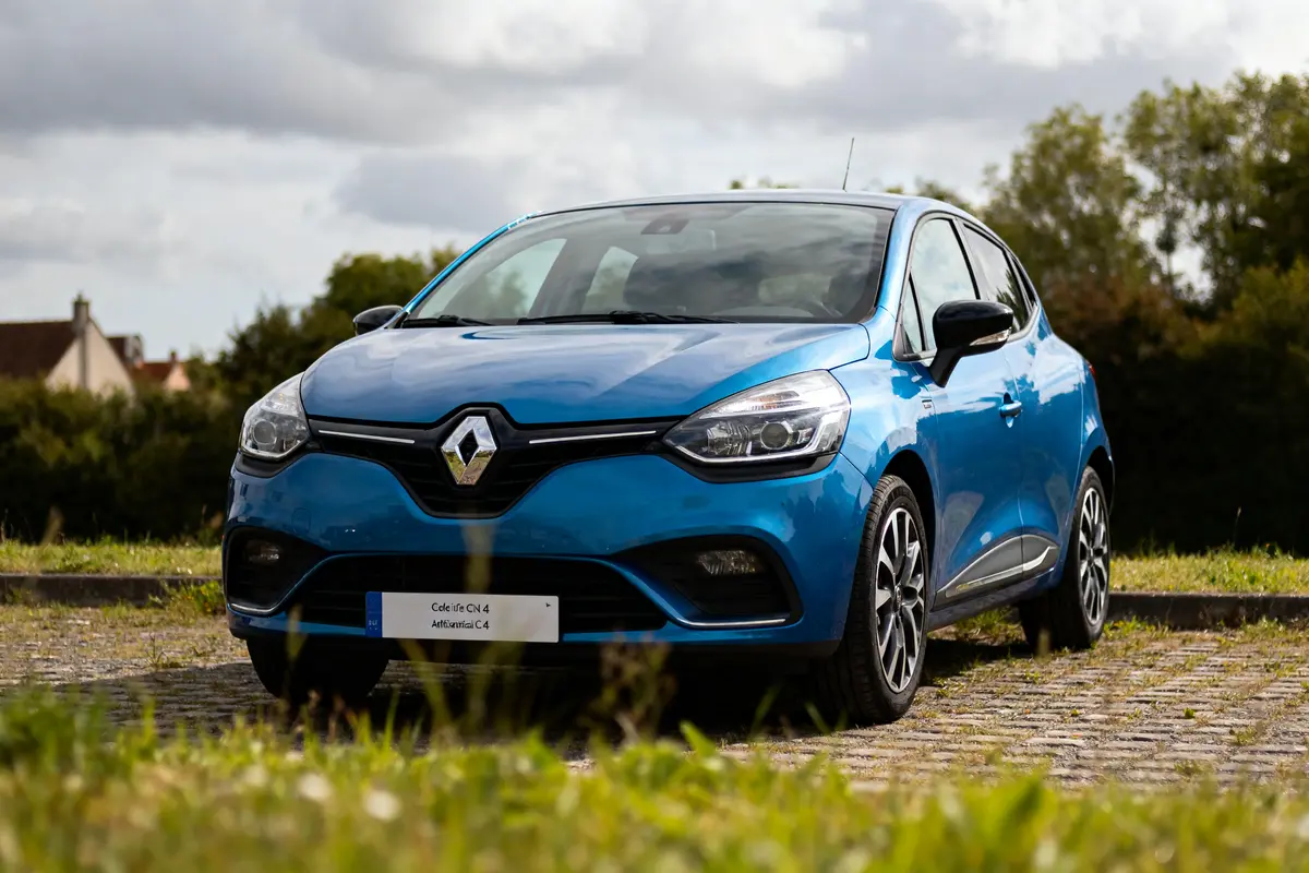 Voiture bleue Renault Clio garée sur route pavée, arrière-plan bucolique, ciel nuageux.