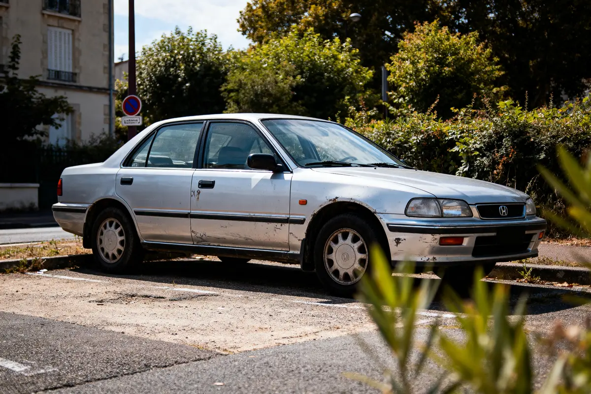Vieille voiture blanche Honda garée sur parking extérieur, devant végétation et bâtiment résidentiel, ensoleillé.