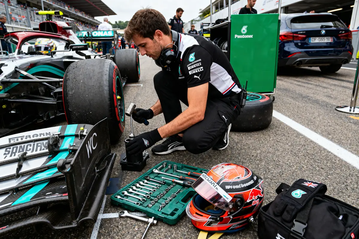 Un mécanicien prépare une voiture de course, outils et casque à proximité, sur la piste d'un circuit.