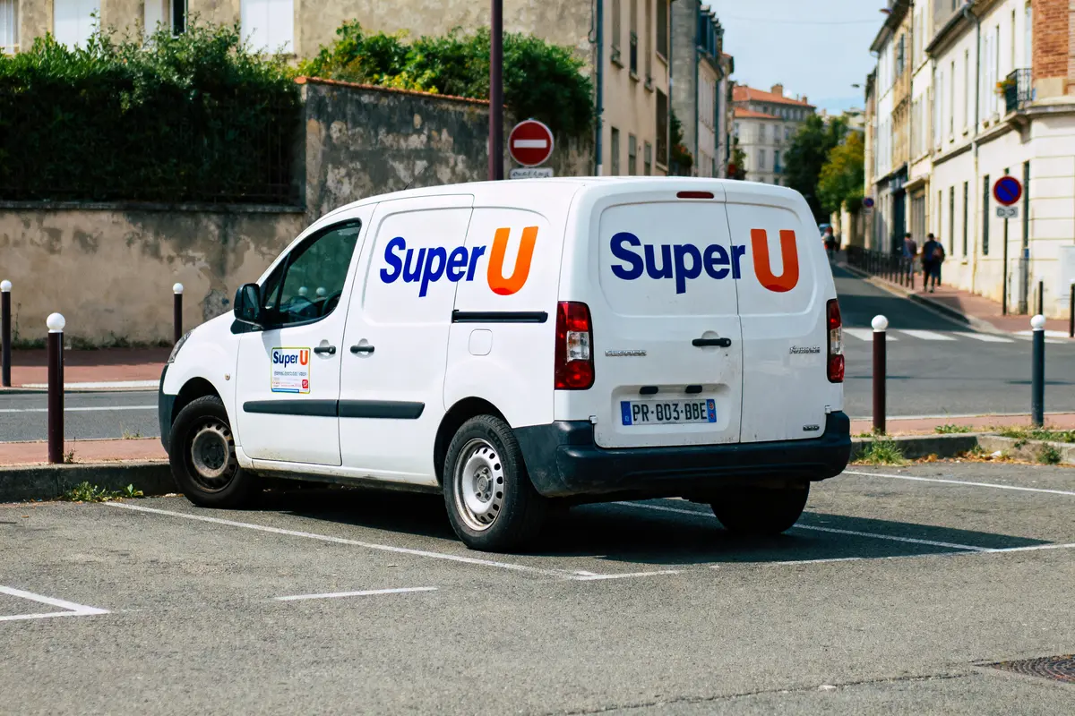 Utilitaire blanc Super U garé dans une rue résidentielle, bâtiments en arrière-plan.