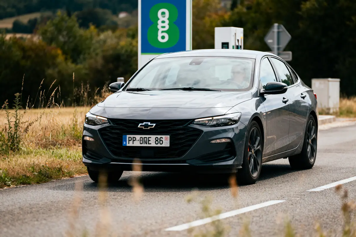 Voiture grise Chevrolet sur route rurale, devant une station de recharge, arrière-plan boisé.