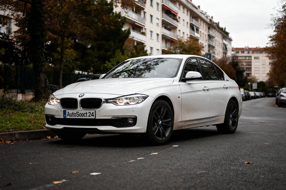 Voiture BMW blanche sur rue bordée d'arbres avec plaque AutoScout24 visible