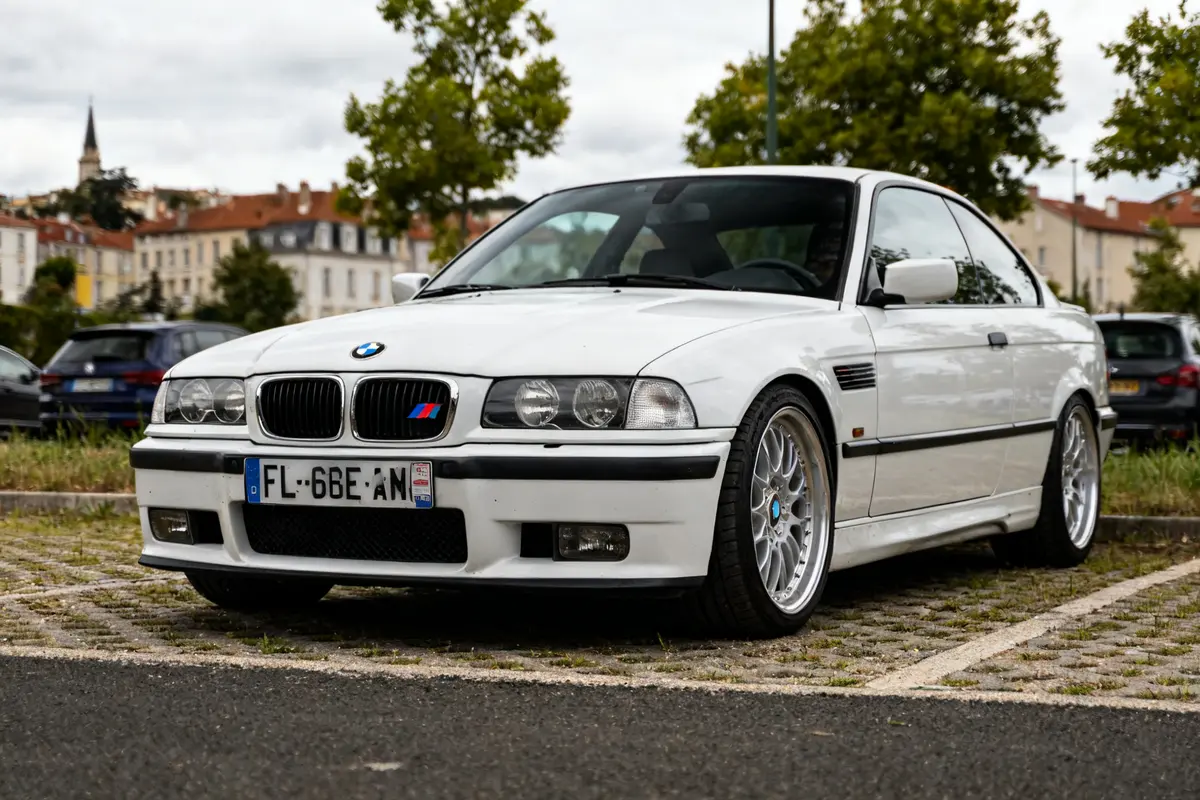 BMW M3 blanche garée sur parking pavé, arbres et bâtiments en arrière-plan