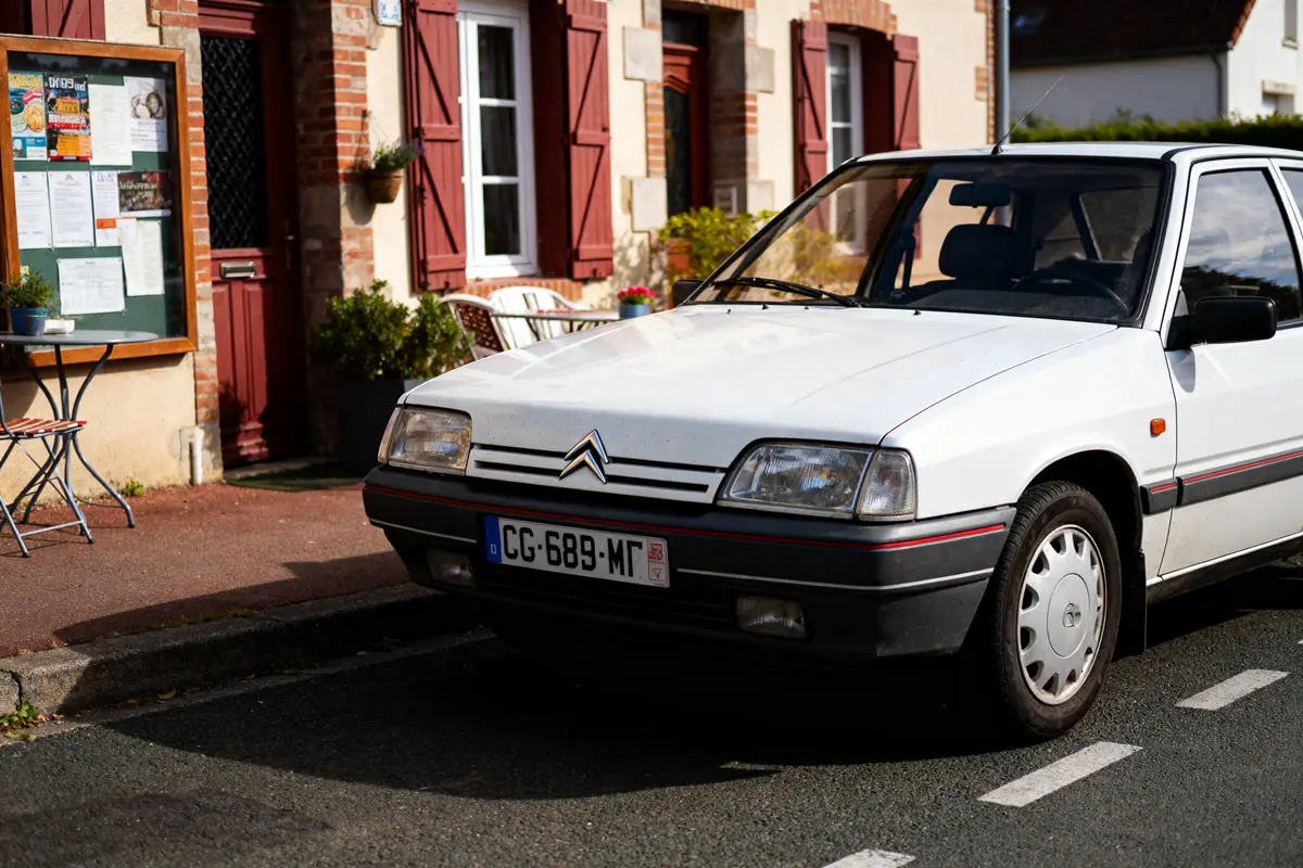 Voiture blanche Citroën BX garée devant une maison avec volets rouges