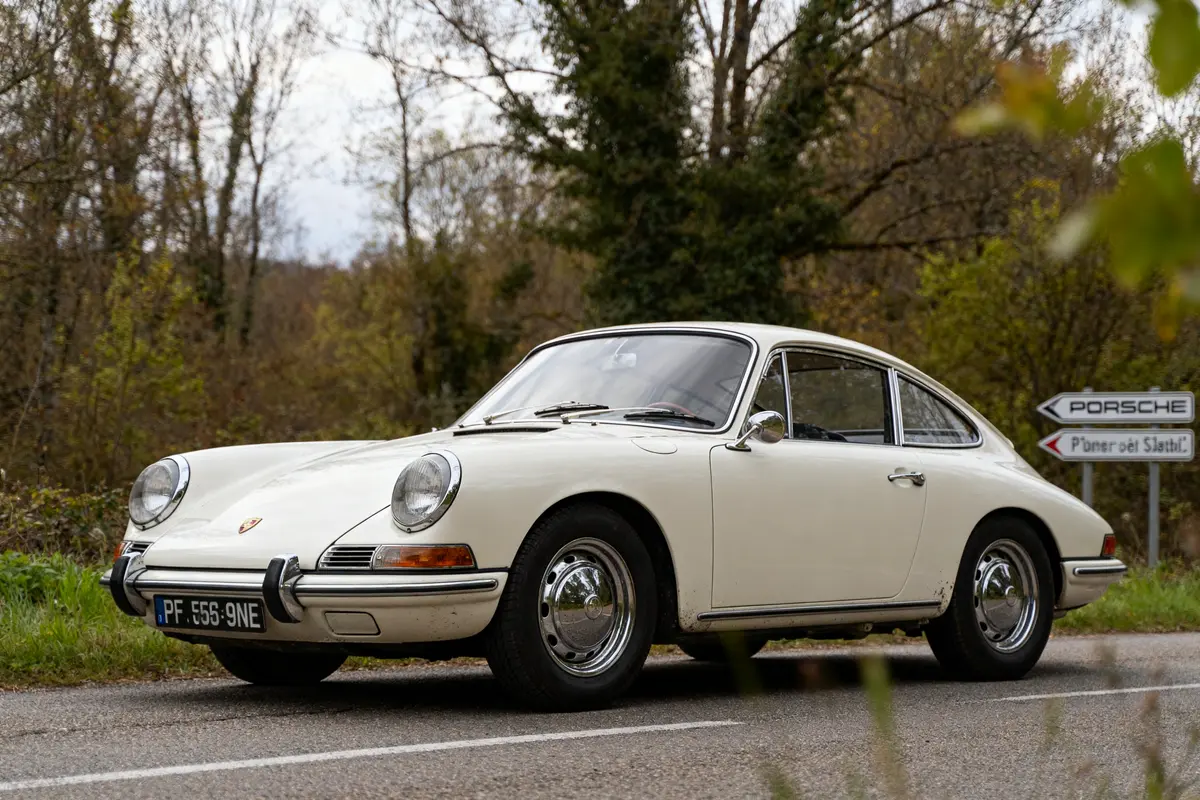 Porsche ancienne blanche garée sur route bordée d'arbres et panneau indicateur Porsche