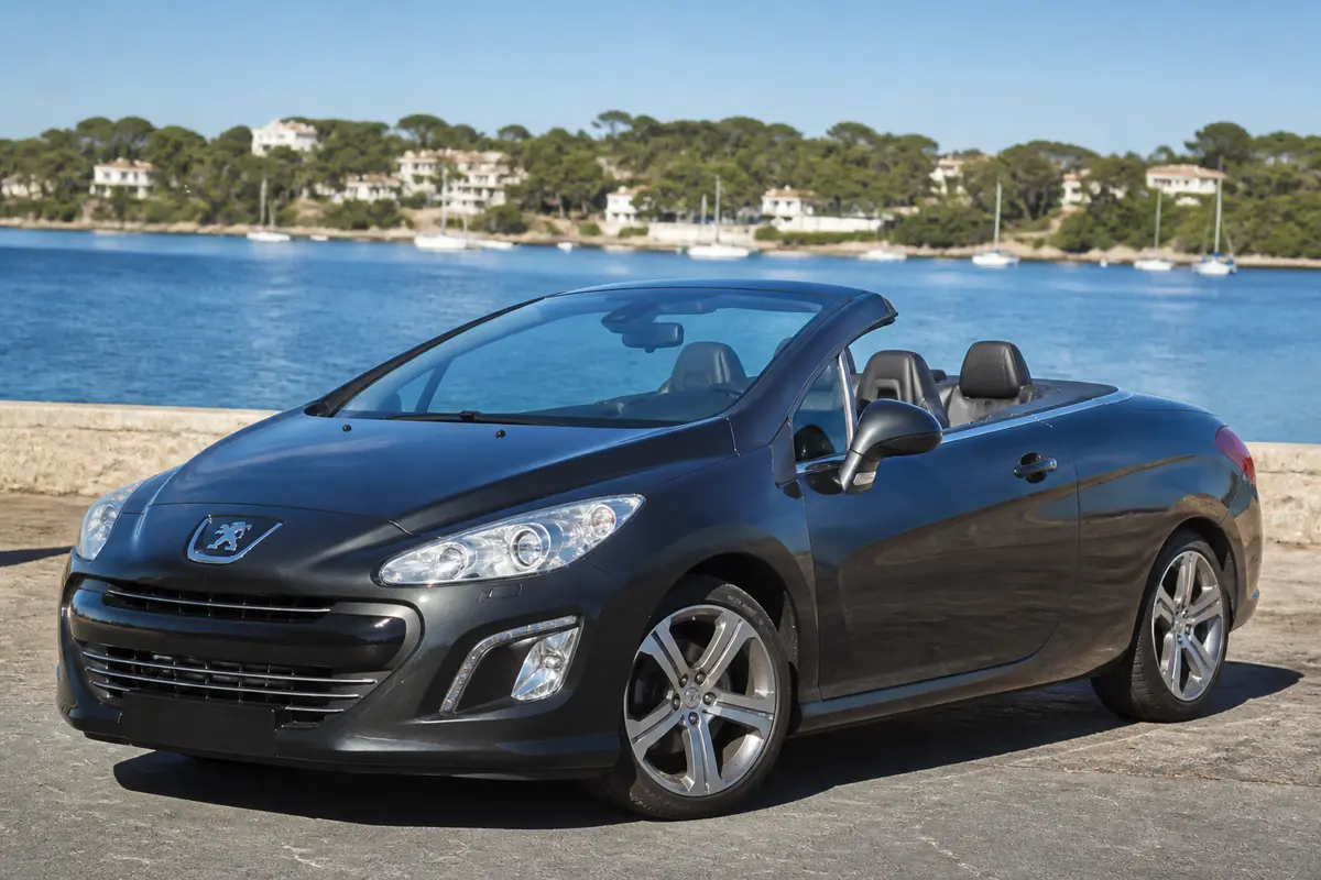 Voiture décapotable Peugeot noire garée près d'un lac, ciel bleu et maisons en arrière-plan.
